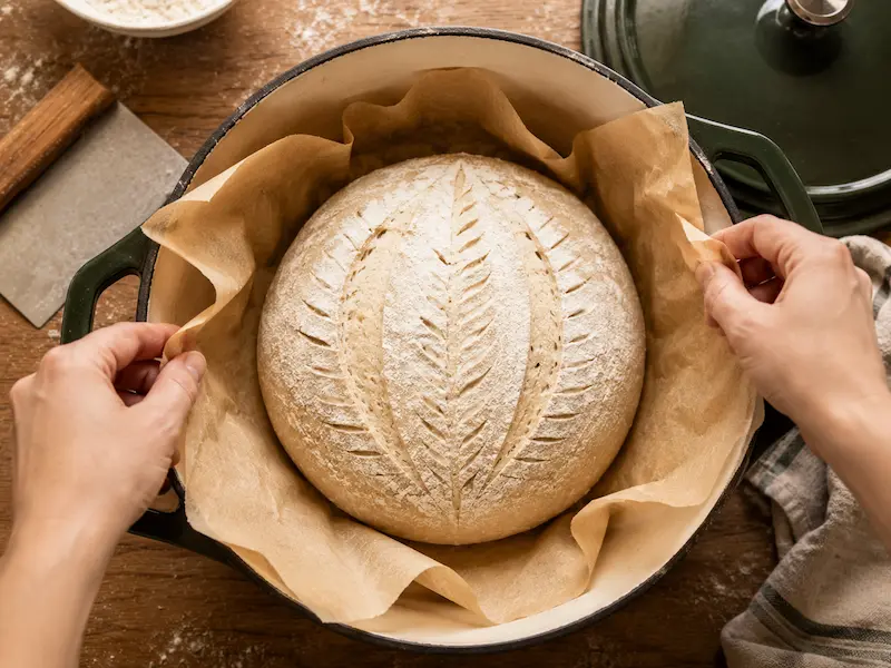 Scored sourdough dough on parchment paper being lowered into a preheated Dutch oven