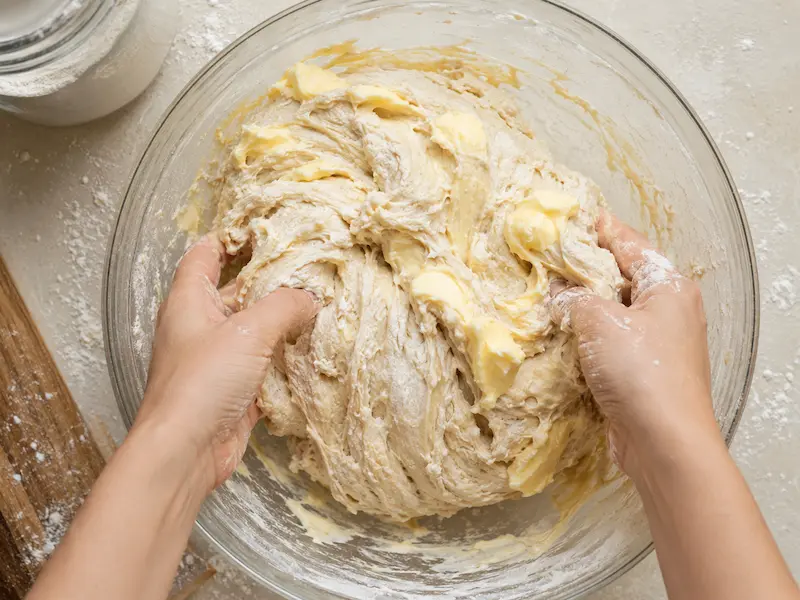 Hands folding softened butter into shaggy sourdough sandwich bread dough in a glass mixing bowl
