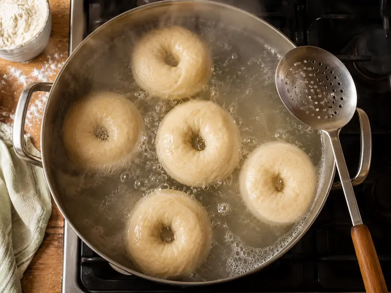 Sourdough bagels boiling and floating in a pot of water with a slotted spoon resting on the edge