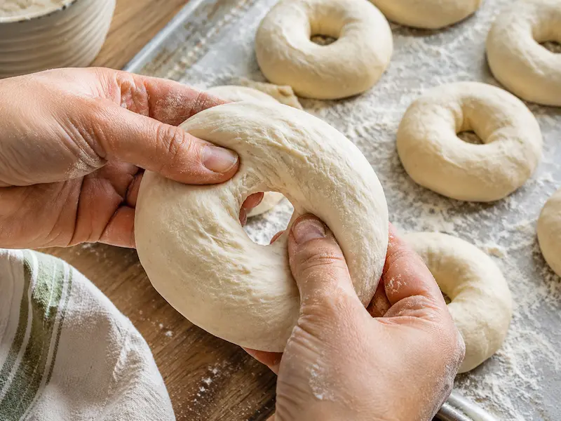 Hands poking and stretching a hole through sourdough bagel dough using the poke method with shaped rings on a tray