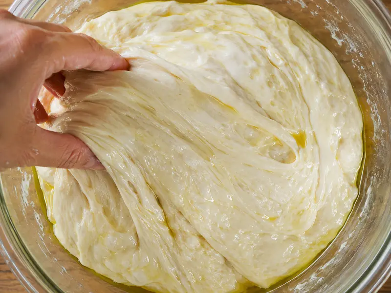 Overhead close up of wet glossy sourdough focaccia dough being folded by hand in a glass bowl