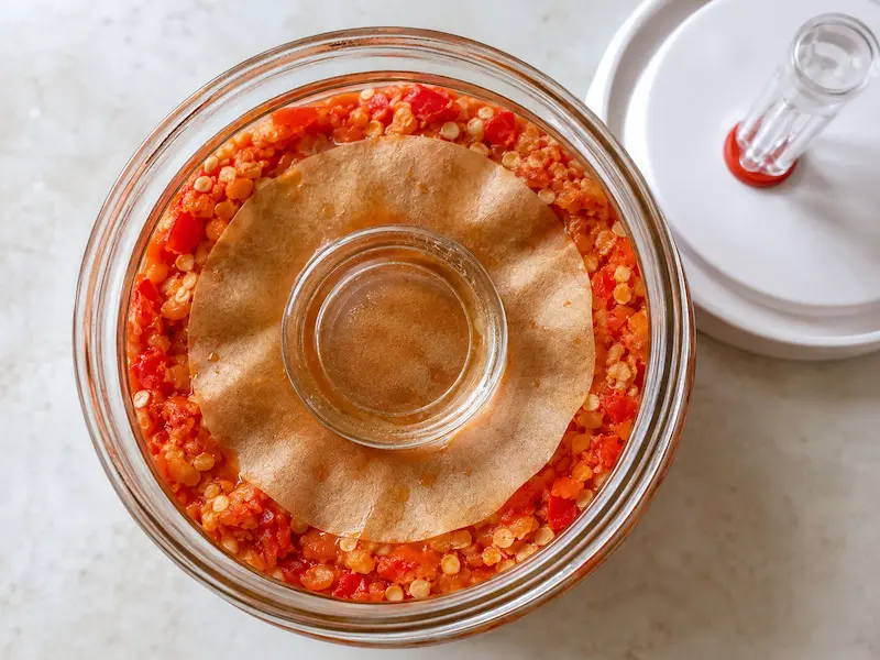 Overhead close up of pepper mash packed into a jar with a parchment cartouche and glass fermentation weight on top