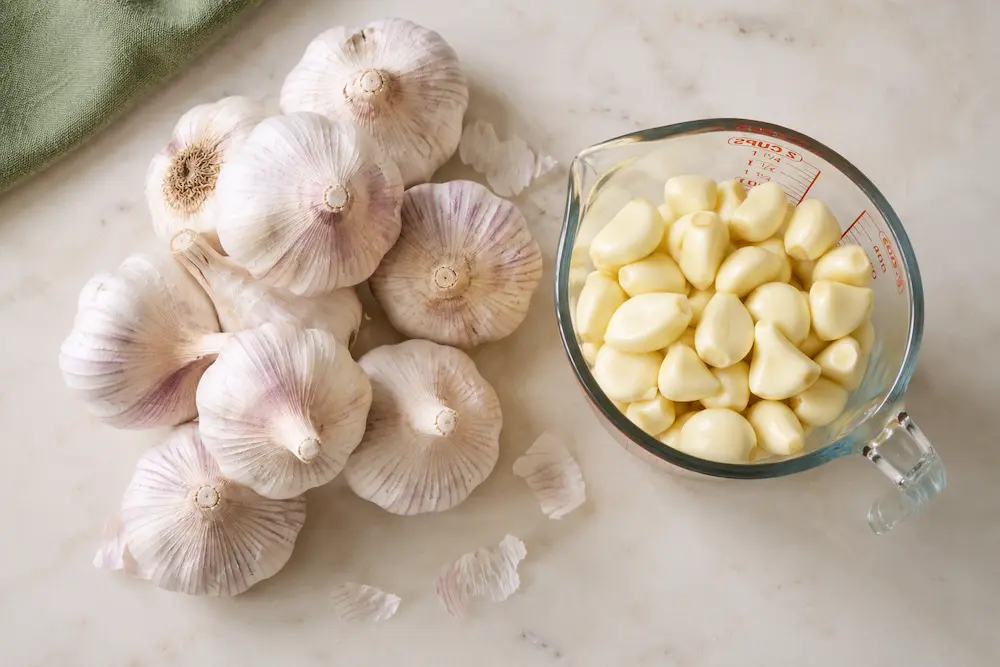 Unpeeled garlic bulbs next to a measuring cup of peeled cloves showing volume comparison