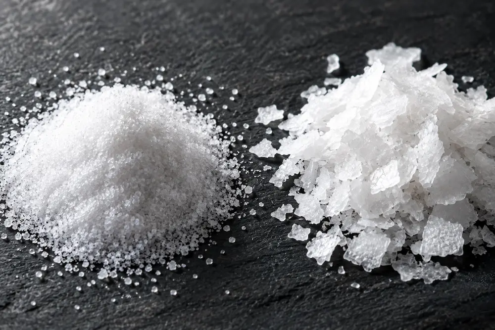 Close-up comparison of fine pickling salt crystals next to coarse kosher salt flakes